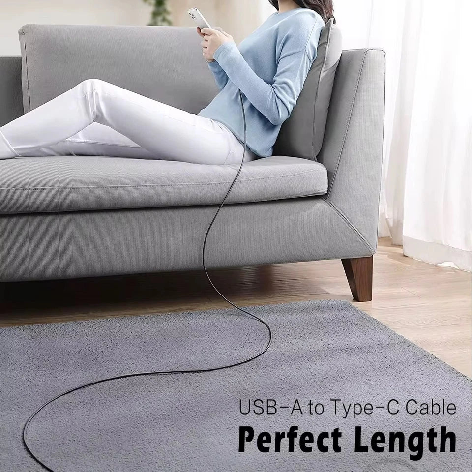 A lifestyle photo of a woman relaxing on a sofa while charging her phone with a long, black USB-A to Type-C cable that reaches across the floor.